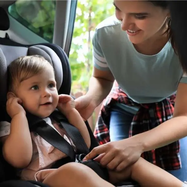 niña pequeña sonriente sentada en asiento isofix, con mujer acomodándole el cinturón